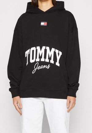 Personne portant un sweat à capuche noir avec le texte "TOMMY Jeans" et logo sur le devant, associé à un pantalon blanc, sur un fond clair uni.