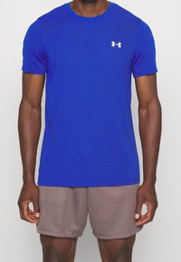 Man i en blå Under Armour träningsskjorta och ljusbruna meshshorts, som står mot en enfärgad bakgrund.