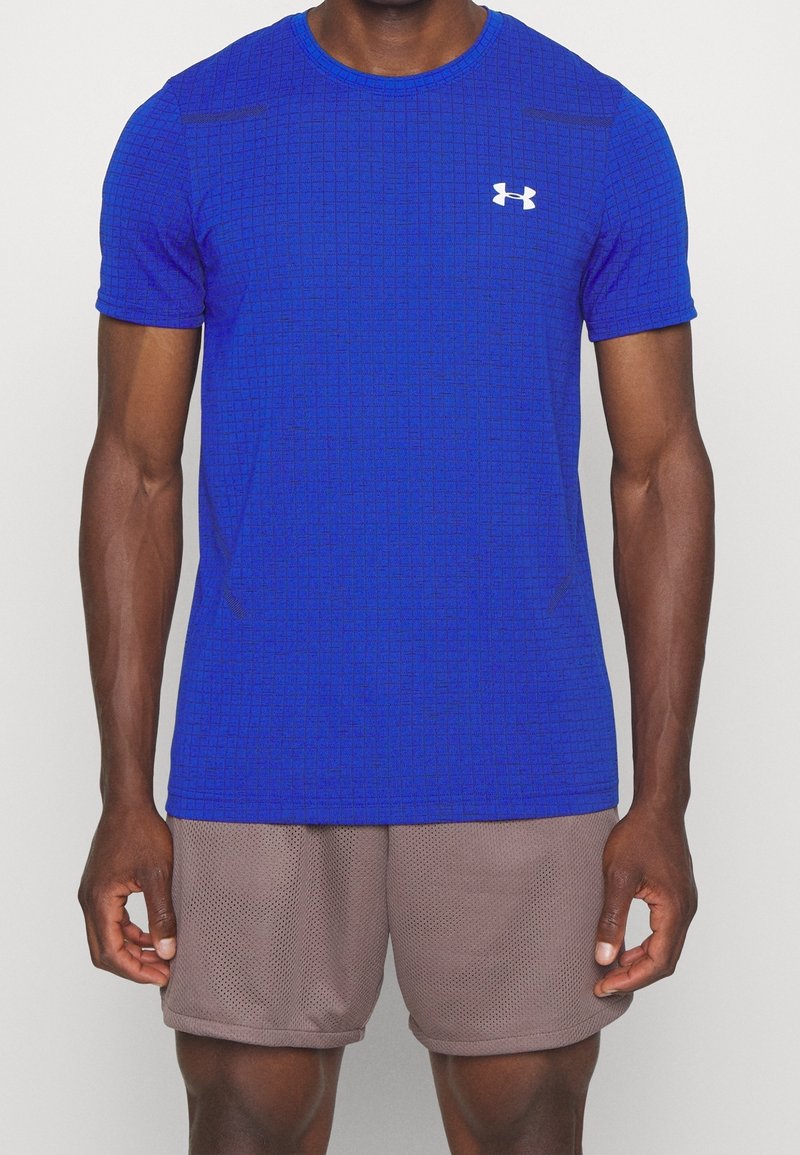 Man i en blå Under Armour träningsskjorta och ljusbruna meshshorts, som står mot en enfärgad bakgrund.