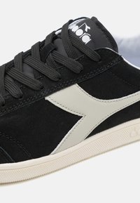 Sneakers in suede nero con una swoosh in pelle color crema, suola in gomma bianca texturizzata e lacci neri con un logo sulla linguetta.