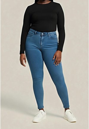 Personne portant un jean bleu bien ajusté, un haut noir à manches longues et des baskets blanches, debout devant un fond uni.