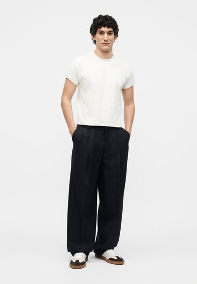 Άνδρας στέκεται με τα χέρια στις τσέπες, φοράει λευκό κοντομάνικο πουκάμισο, μαύρο παντελόνι με φαρδιά μπατζάκια και λευκά αθλητικά παπούτσια με καφέ σόλες.