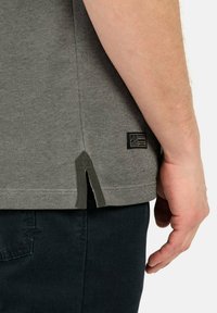 Graues, strukturiertes T-Shirt mit seitlichen Schlitzen, kontrastierenden grünen Akzenten am Saum und einem kleinen schwarzen Etikett in der unteren rechten Ecke, kombiniert mit dunklen Hosen.