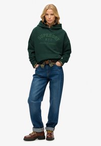 Sudadera con capucha de color verde oscuro y bolsillo frontal, con el texto "SUPERDRY N.Y.C" en un tono verde más claro, llevada sobre una camisa a cuadros y jeans azules. Botas marrones con cordones.