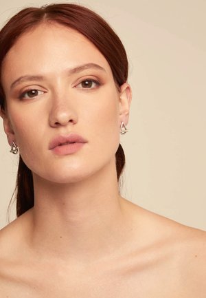 Pendientes geométricos plateados con un diseño triangular y un acento circular. Textura suave sobre un fondo de tono de piel neutro.