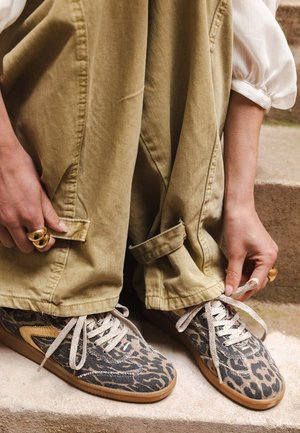 Femme en train de lacer des baskets imprimées léopard, portant un pantalon beige large et un blouse blanche à manches bouffantes, assise sur des marches en pierre.