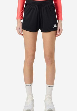 Mujer vestida con pantalones cortos deportivos negros de Kappa, calcetines blancos de Kappa, zapatillas blancas y una camiseta de manga larga roja, de pie frente a un fondo plano.