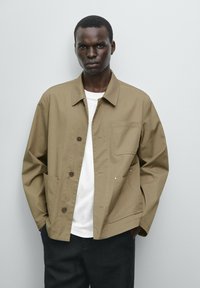 Khaki katoenen jack met een knoopsluiting, verborgen zakken en een borstzak. Draag het over een wit shirt, gecombineerd met donkere broeken.
