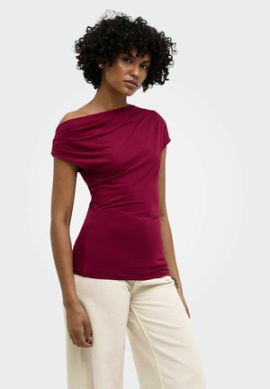 Femme aux cheveux bouclés portant un top bordeaux à une épaule et un pantalon beige sur un fond clair uni.