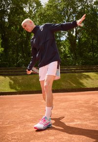Joueur de tennis portant une veste marine, un short rose clair avec des accents turquoise et des chaussures de sport blanches avec des accents roses, se préparant à servir sur un court en terre battue.