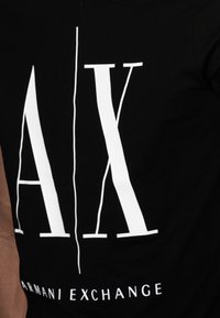 Camiseta de algodón negra con letras blancas en negrita "A/X" y el texto "ARMANI EXCHANGE" en la parte inferior, con una textura suave y un ajuste casual.