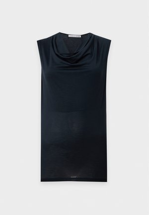 Une femme porte un haut noir sans manches au décolleté drapé, un pantalon à jambes larges et des escarpins pointus, sur un fond de studio clair et uni.