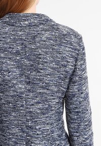 Detailní záběr na záda osoby oblečené ve svetru s modro-bílou texturou a vysokým límcem, na jednobarevném pozadí.