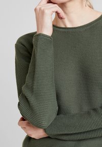 Maglione lavorato a maglia verde scuro con un motivo testurizzato, scollatura tonda e maniche lunghe. Il tessuto sembra spesso e caldo. Le mani sono appoggiate in modo pensieroso.
