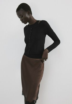 Femme ajustant une jupe en satin marron avec ourlet en dentelle, portant un haut noir à manches longues boutonné, sur fond blanc uni.