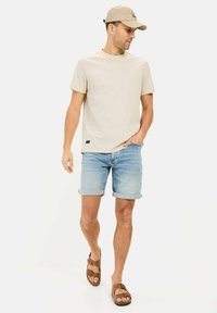 T-shirt en coton beige, shorts en denim bleu, sandales brunes. Le t-shirt a un col rond. La tenue est décontractée et estivale.