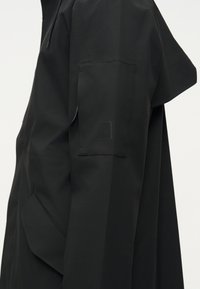 Κοντινό του μανικιού μαύρου μπουφάν με ορθογώνια τσέπη και διακριτικό κεντημένο λογότυπο της μάρκας στο πάνω μέρος του χεριού.