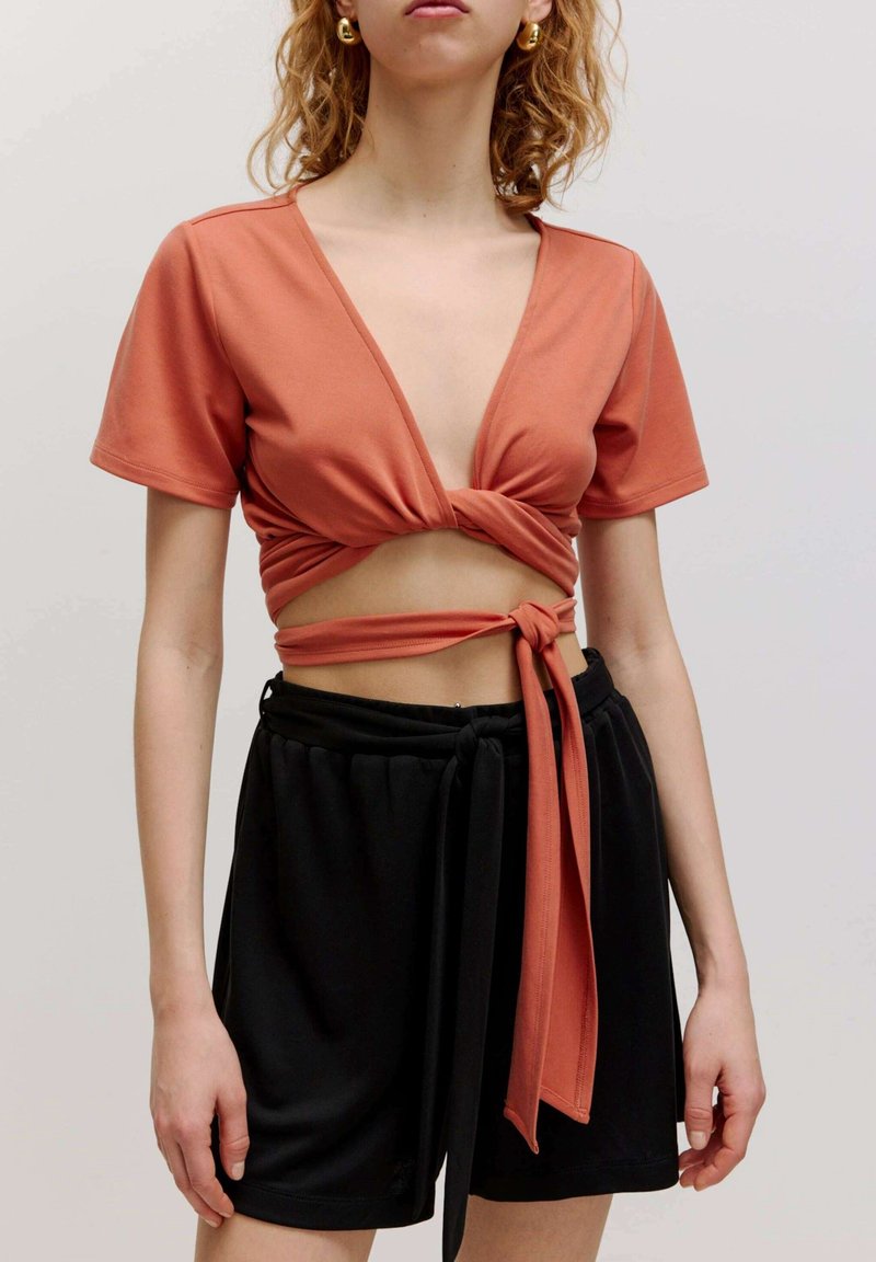 Femme portant un crop top portefeuille couleur rouille à manches courtes et un short noir taille haute noué à la taille avec des ceintures en tissu assorties.