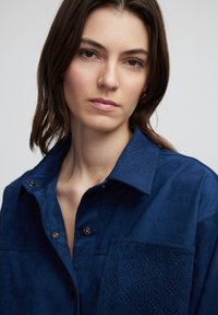 Marineblauw suède shirt met een button-down kraag, voorzien van een geweven zak met een geometrisch patroon en metalen knopen.