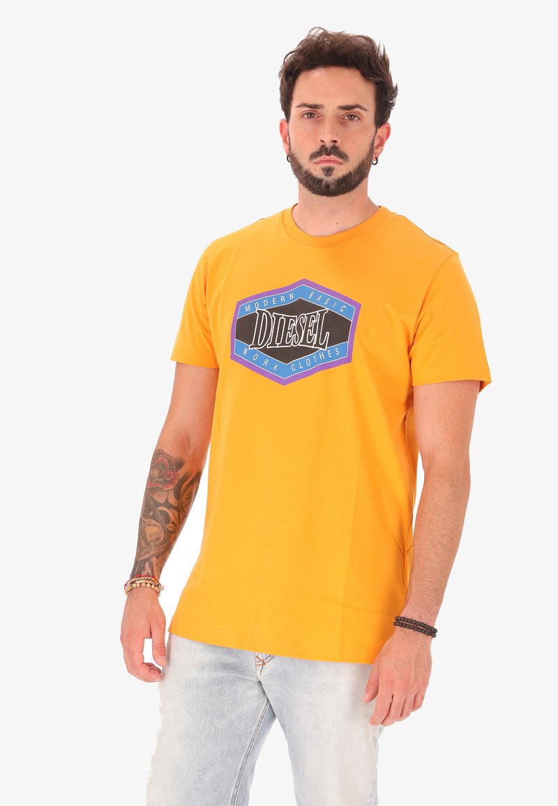 Man met een baard en tatoeages draagt een oranje Diesel T-shirt en lichtgewassen jeans, staand tegen een effen witte achtergrond.