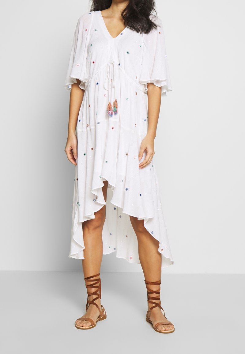 Femme portant une robe blanche longue jusqu'aux chevilles avec des manches volantes et de petits motifs à pois colorés, associée à des sandales marron à lacets.