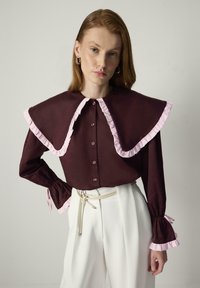 Bordeauxblouse met grote, roze gerimpelde kraag en mouwen; heeft knopen aan de voorkant en aansluitende boorden. Gecombineerd met hoogtaillles witte broek.