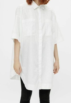 Personne portant une chemise blanche à manches courtes surdimensionnée avec deux poches poitrine et un pantalon noir.