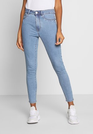 Jeans ajustados de denim azul claro, con cintura alta, diseño de cinco bolsillos y costuras tradicionales. Combinados con zapatillas blancas.