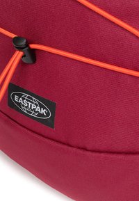 Punainen kangaskassi, jossa on oranssit joustoköydet ja musta Eastpak-logotunnus. Tekstuurin näyttää olevan sileä ja siinä on vahva ompelupisto.
