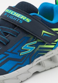 Les baskets bleues et marines présentent un dessus en tissu texturé avec des accents vert fluo, une sangle Velcro et une semelle blanche rembourrée.