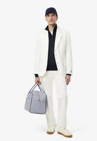 Witte blazer over een marineblauwe ritssluiting-top, gecombineerd met witte broek. Hij houdt een grijze handtas met dubbele bovenhandvatten en een ritsluiting vast. Tan schoenen.