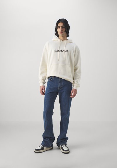 Cremefarvet hoodie med frontlomme og grafisk detalje, parret med blå jeans og sort-hvide sneakers. Glat tekstur og afslappet pasform.
