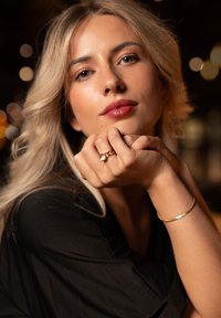 Zelta gredzens ar apļveida akmeni, ko valkā uz rokas ar zelta aproci. Modelis ar viļņainiem blondiem matiem un melnu blūzi, pozējot ar rokām tuvumā sejas.