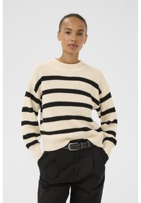 Cream sweater with black horizontal stripes, ribbed texture, and a high neckline, paired with black trousers and a belt.