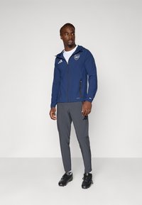 Marine windbreaker met rits, voorzien van een wit logo en drie strepen. Gecombineerd met grijze broek en zwarte sportschoenen. Glad stof, aansluitend ontwerp.
