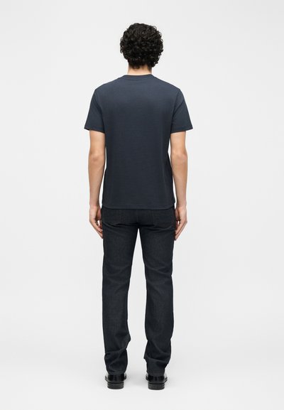 Άνδρας με σγουρά μαλλιά φορώντας ένα μονόχρωμο σκούρο κοντομάνικο μπλουζάκι, σκουρόχρωμο τζιν και μαύρα παπούτσια, στέκεται με την πλάτη προς το μέρος του θεατή σε λευκό φόντο.