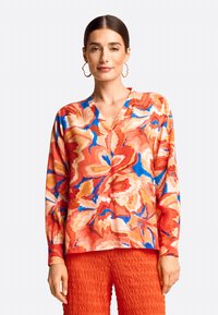 Langärmlige V-Ausschnitt-Bluse aus fließendem Stoff. Mit einem lebhaften Blumenmuster in Rot-, Blau- und Orangetönen. Kombiniert mit strukturierten roten Hosen.