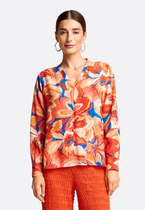 Langärmlige V-Ausschnitt-Bluse aus fließendem Stoff. Mit einem lebhaften Blumenmuster in Rot-, Blau- und Orangetönen. Kombiniert mit strukturierten roten Hosen.