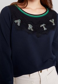 Tmavě modrá mikina s límcem se zeleno-zlatými pruhy, s krajkovým panelem a korálkovým detailem "ARTY" na přední straně. Materiál ze směsi bavlny.