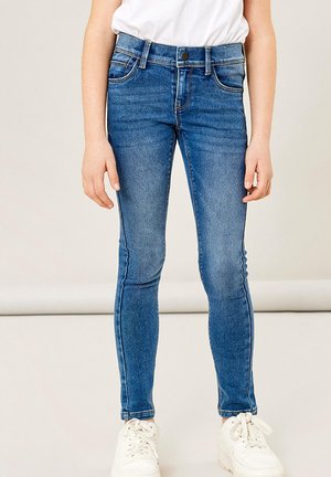 Kind trägt blaue Skinny-Jeans mit Fronttaschen und weiße Turnschuhe, steht vor einem schlichten hellen Hintergrund.