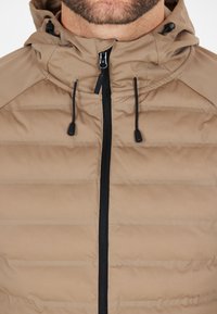 Giacca puffer beige con cappuccio, dotata di chiusura con zip nera e cordini, motivo trapuntato e una texture liscia.