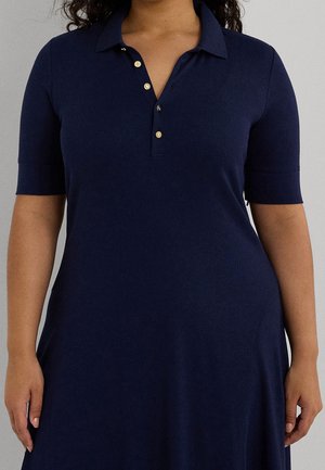 Frau trägt ein marineblaues Polokleid mit kurzen Ärmeln und goldenen Knöpfen, steht entspannt mit den Händen an den Seiten vor einem grauen Hintergrund.