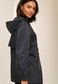 Zwarte nylon regenjack met capuchon, ritssluiting en elastische manchetten. Beschikt over een voorzak met flap en een iets ontspannen pasvorm.
