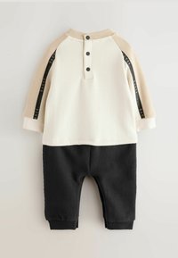 Haut à manches longues blanc et beige avec trois boutons noirs et des bandes latérales noires, associé à un pantalon noir avec des poignets élastiques.