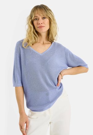 Mujer rubia con cabello ondulado, vestida con un suéter de punto de manga corta color lila claro y pantalones blancos, posando con una mano en la cadera.