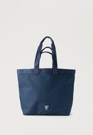 Tamnoplavna Lacoste torba s dvojno ročico in logotipom na sprednji strani, enobarvno ozadje.