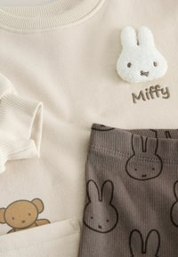 Beżowa bluzka z prążkowanym dekoltem, z aplikacją w postaci puszystego białego króliczka oznaczonego napisem "Miffy". Brązowe paskowane spodnie z nadrukiem króliczych twarzy.