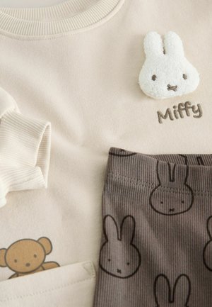 Beżowa bluzka z prążkowanym dekoltem, z aplikacją w postaci puszystego białego króliczka oznaczonego napisem "Miffy". Brązowe paskowane spodnie z nadrukiem króliczych twarzy.