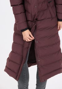 Bordeaux pufferjakke med et langt, quiltet design, der har lynlås foran og trykknapper, sammen med grå jeans og en sort manschet.