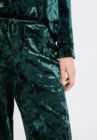 Zelená velurová souprava na domácí nošení s elastickým pasem a dlouhými rukávy. Materiál má měkkou, lesklou texturu s mírným leskem.